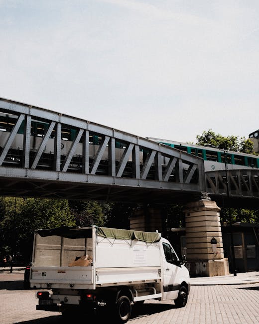 A white moving van with an open cargo area parked on a paved street beneath an elevated railway bridge with green and white trains passing overhead. The van is positioned near a building entrance or l